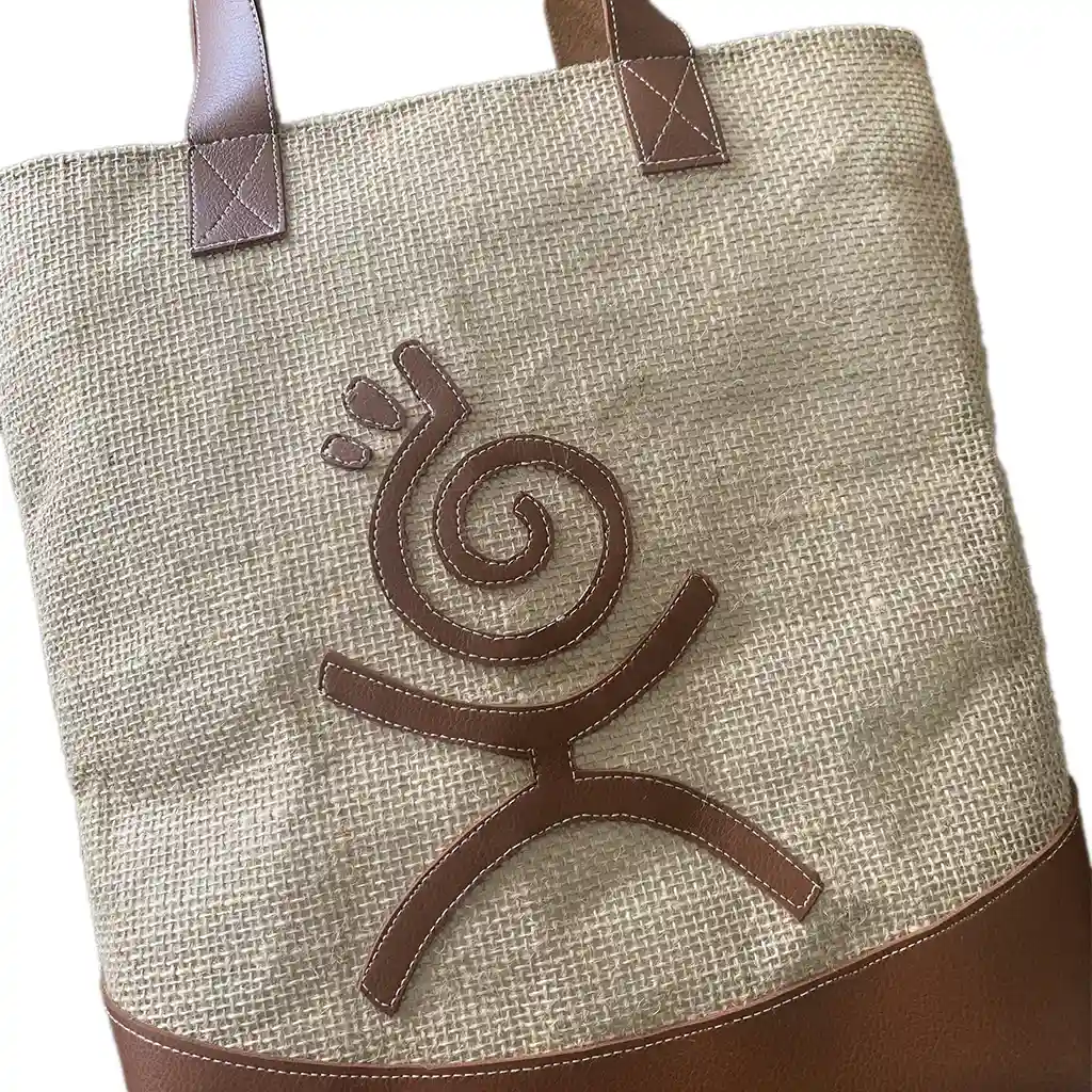 Sac cabas KYDRO en toile de jute beige et simili-cuir camel, motif spiralé marron, sur fond orangé façon coucher de soleil antillais.
