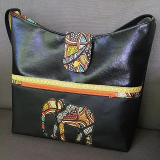 Sac cabas noir en simili cuir avec fermeture à rabat, orné d’un éléphant en tissu wax multicolore et d’un zip orange contrastant.